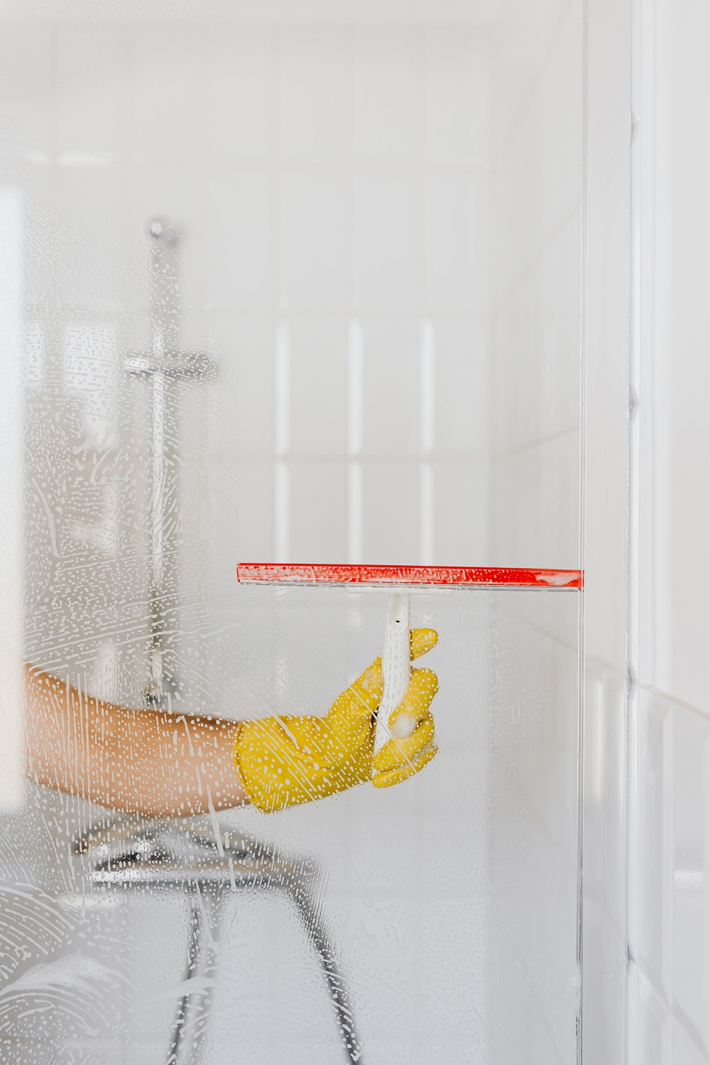 Biely ocot pomáha žene pri umývaní skla sprchovacieho kúta od vodného kameňa. Žena stiera kvapky vody stierkou v rukaviciach.